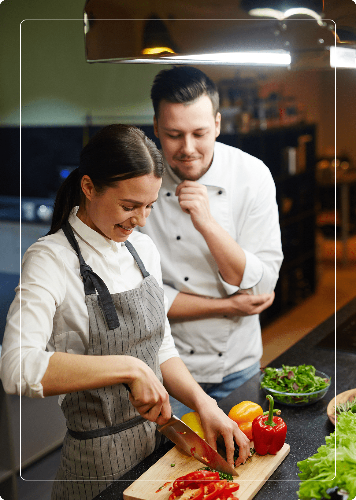 Two chefs in a kitchen; a woman slices a red bell pepper on a cutting board while a man watches. Various vegetables are on the counter.