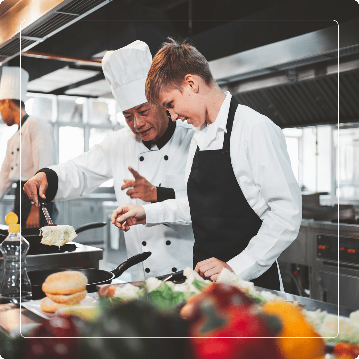 Group 2 (5) Chef instructs young cook as they prepare food together in a commercial kitchen, with vegetables and kitchenware visible in the foreground.