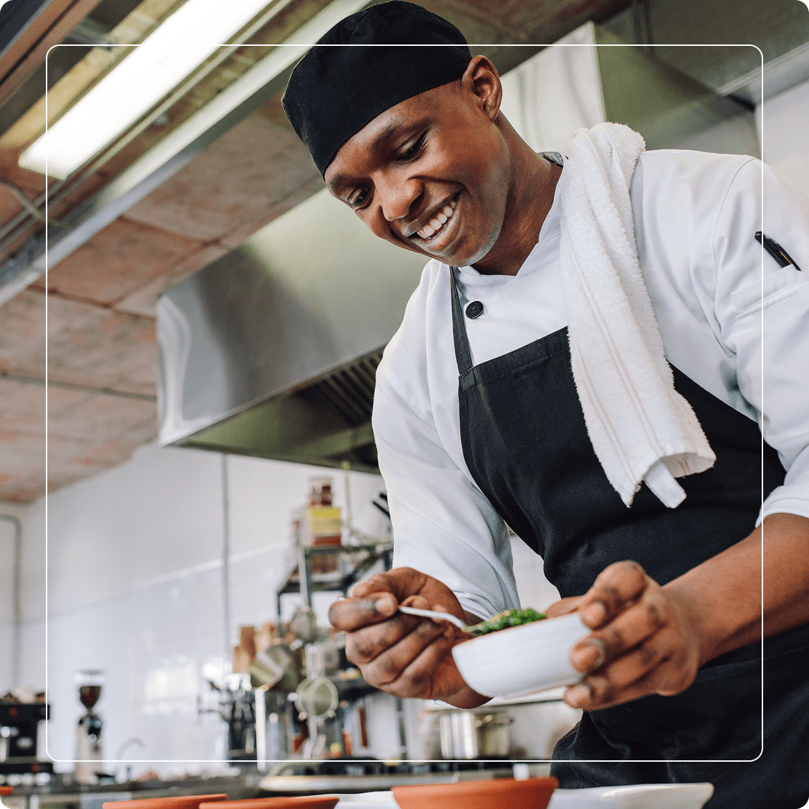 Group 2 (6) Chef in a black apron and hat smiling while garnishing a dish with greens in a professional kitchen.