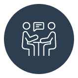 Icon showing two stick figures sitting at a table, facing each other, with a speech bubble between them, enclosed in a circular dark background.