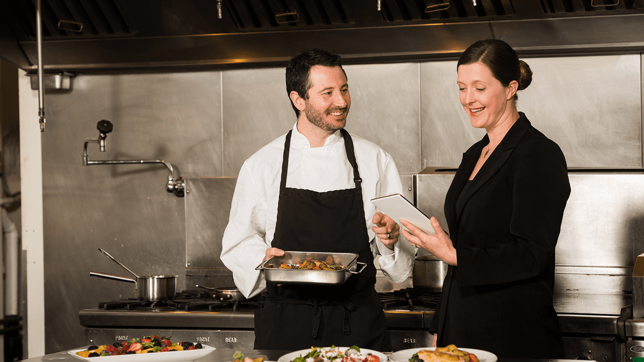 Chef in a kitchen, holding a tray, smiling at a woman in business attire who is holding a tablet. Plates of food are on the counter.