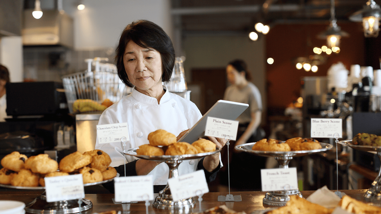 A woman in a chef's coat examines pastries on display in a bakery. There are various scones labeled with flavors like chocolate chip and plain. Another person works in the background.
